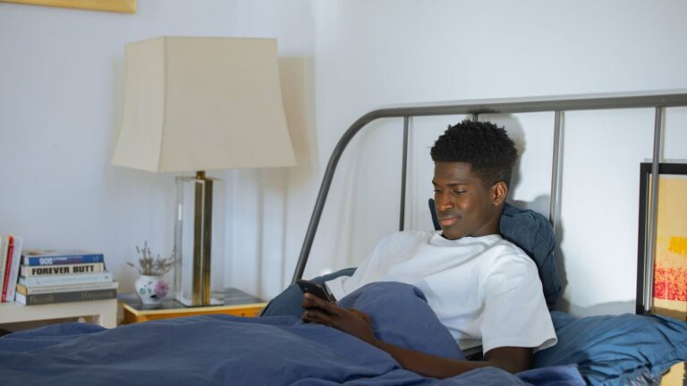 Man in a white shirt sitting in bed using a smartphone beside a lamp