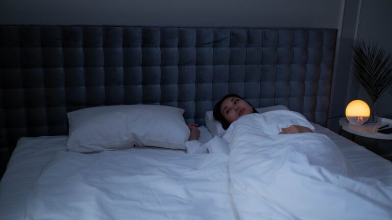 Woman lying awake in a dim bedroom at night, looking up from under white bedding
