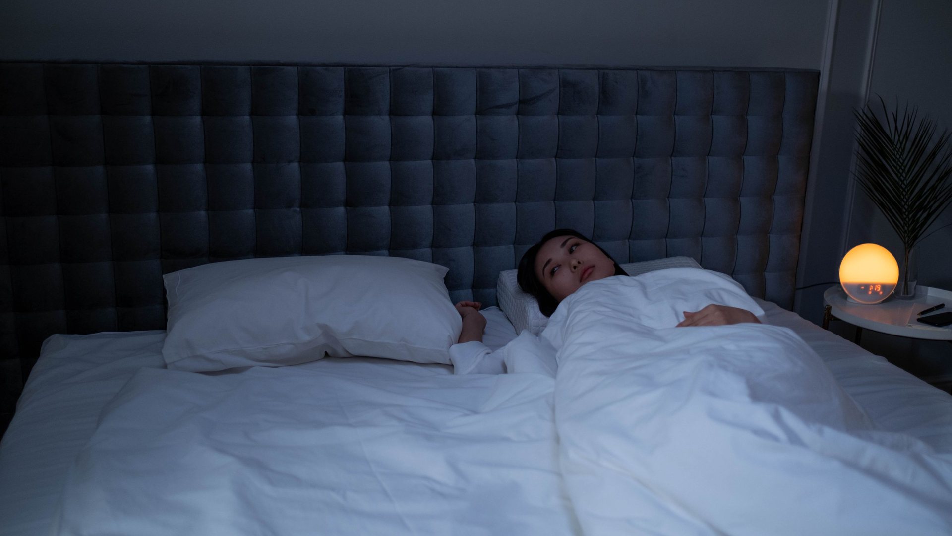 Woman lying awake in a dim bedroom at night, looking up from under white bedding