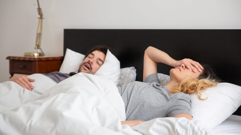 Woman with her hand on her forehead lying awake next to a snoring man