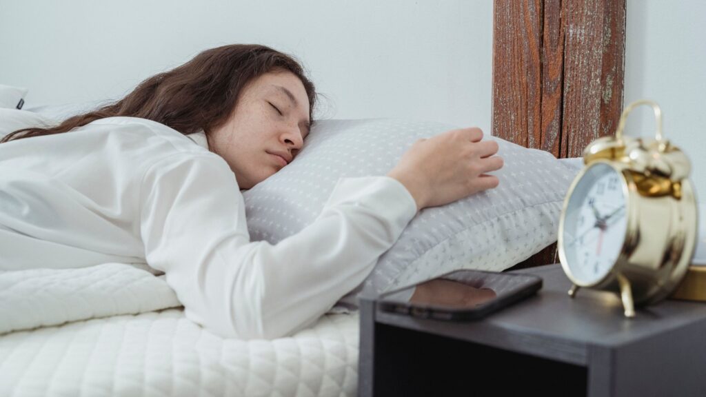 Woman sleeping on her side in white bedding with an alarm clock and phone on the nightstand